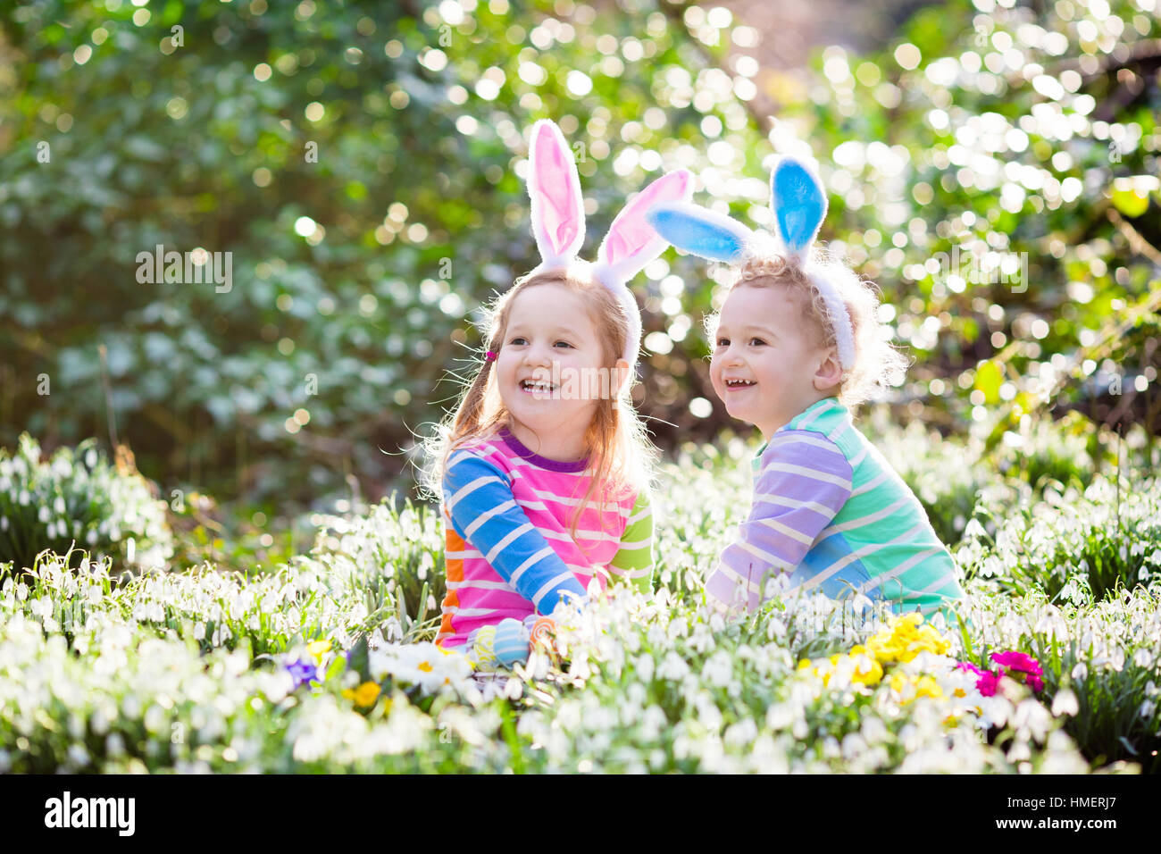 Kids on Easter egg hunt in blooming spring garden. Children with bunny ears searching for colorful eggs in snow drop flowers Stock Photo