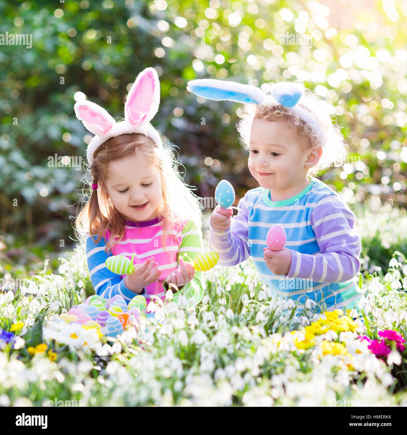 Kids on Easter egg hunt in blooming spring garden. Children with bunny ears searching for colorful eggs in snow drop flowers Stock Photo