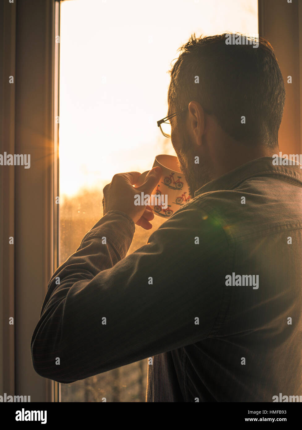 Man drinks from a mug, as warm morning sunlight beams through a window. Stock Photo