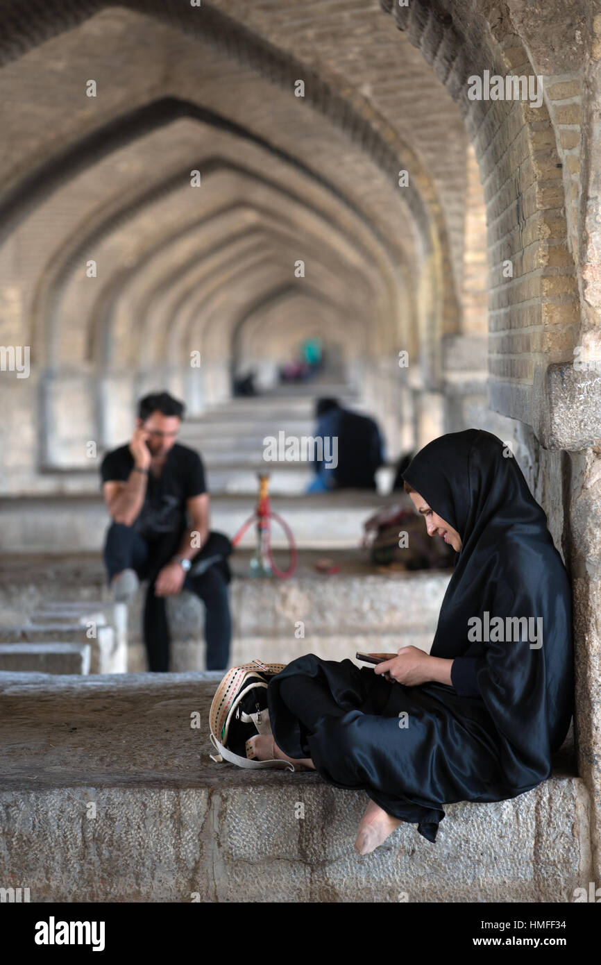 A veiled woman watching hersmart phone under the arches of Si-o-seh Pol Bridge, Isfahan, Iran Stock Photo