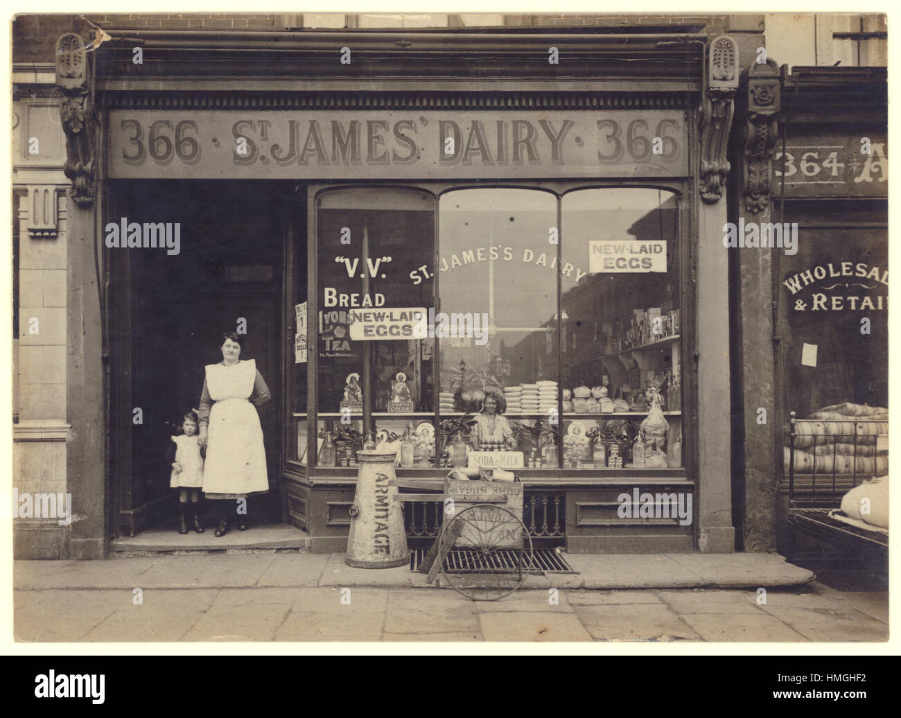 Original early 1900's WW1 era postcard of St. James' Dairy shop / grocery store with female proprietor / owner or staff / assistant and her child, wonderful selection of brands such as Nevill's bread, Tate cube sugar, Armitage, Lyons cocoa and tea. New laid eggs sold loose. London, U.K. circa 1915 Stock Photo