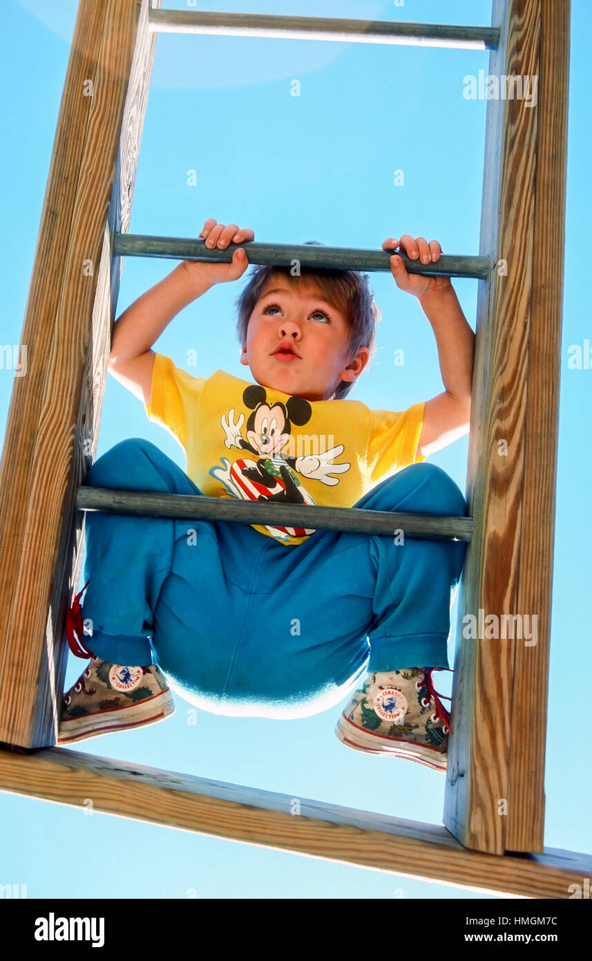 A three year old boy has a worried look on his face as he climbs across a horizontal exercise ladder at a playground in Plainfield, New Hampshire, Uni Stock Photo