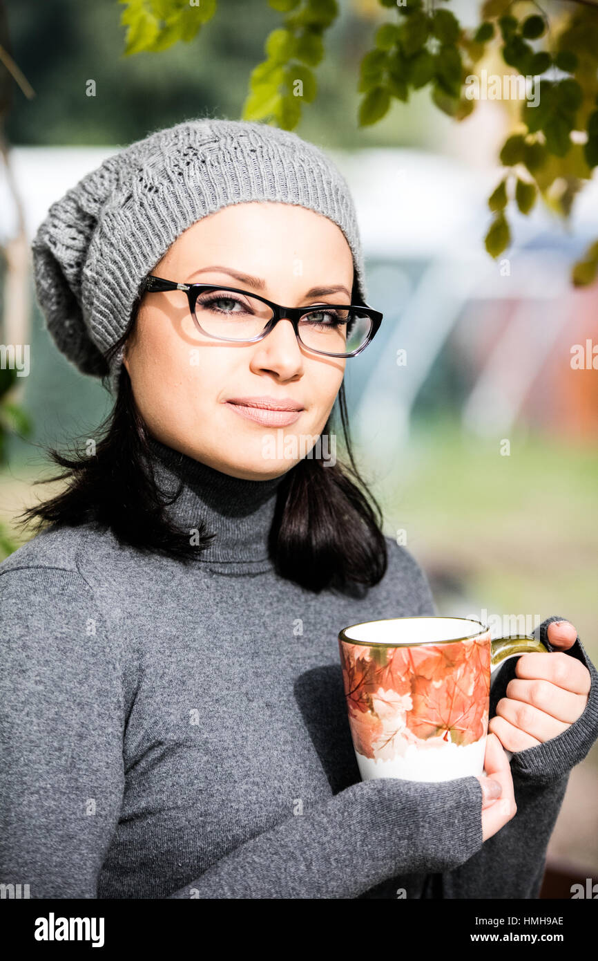 Beautiful green eyed young woman in warm clothes, vision glasses and gray hat smiling, drinking tea in a big cup outdoors in autumn time Stock Photo