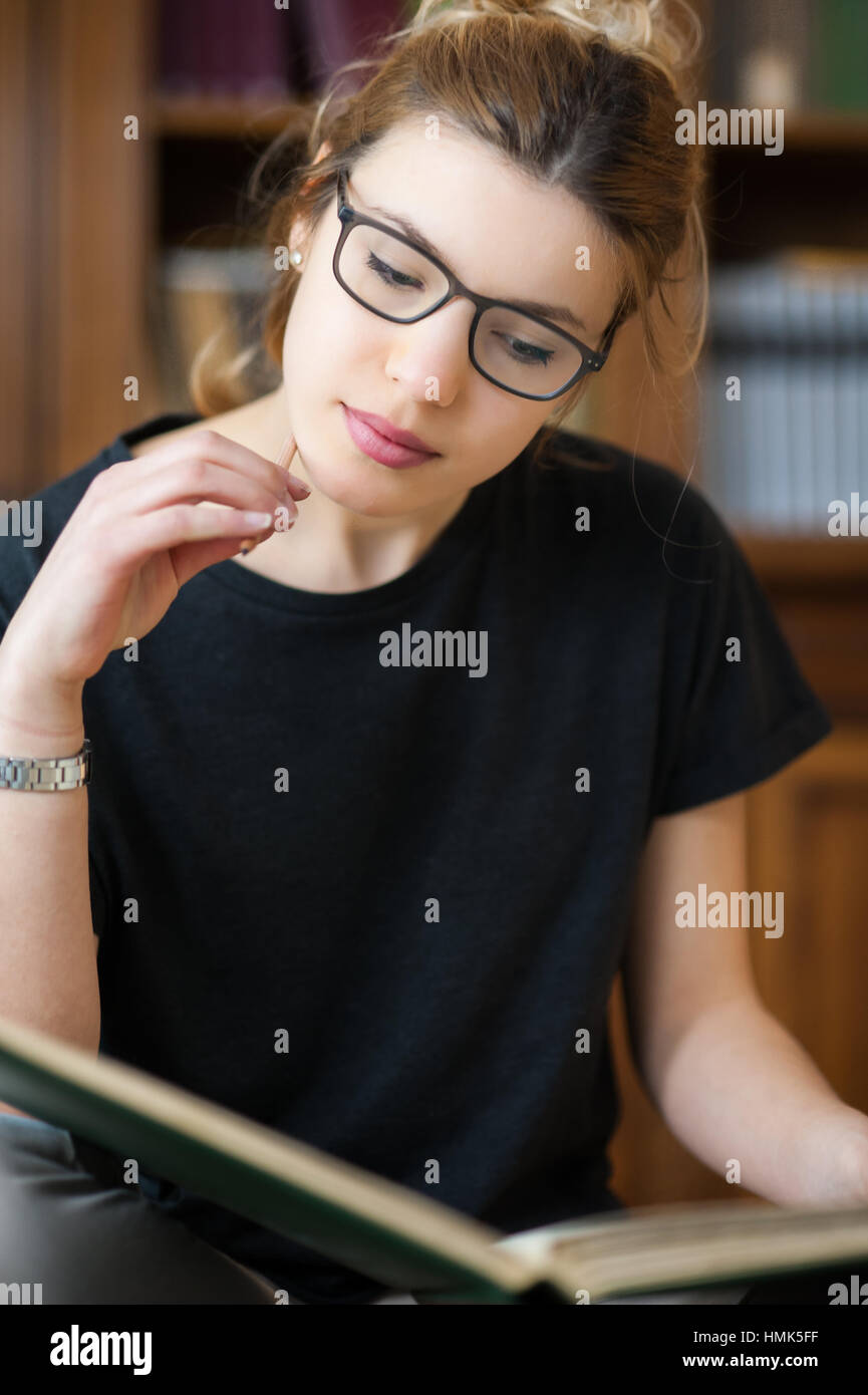 Young female student working in library, focused on book, bookcase in background Stock Photo