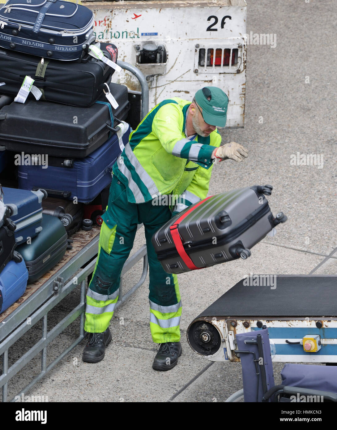 man wearing yellow high-viz jacket loading bags from a trolley onto a baggage-belt truck Stock Photo