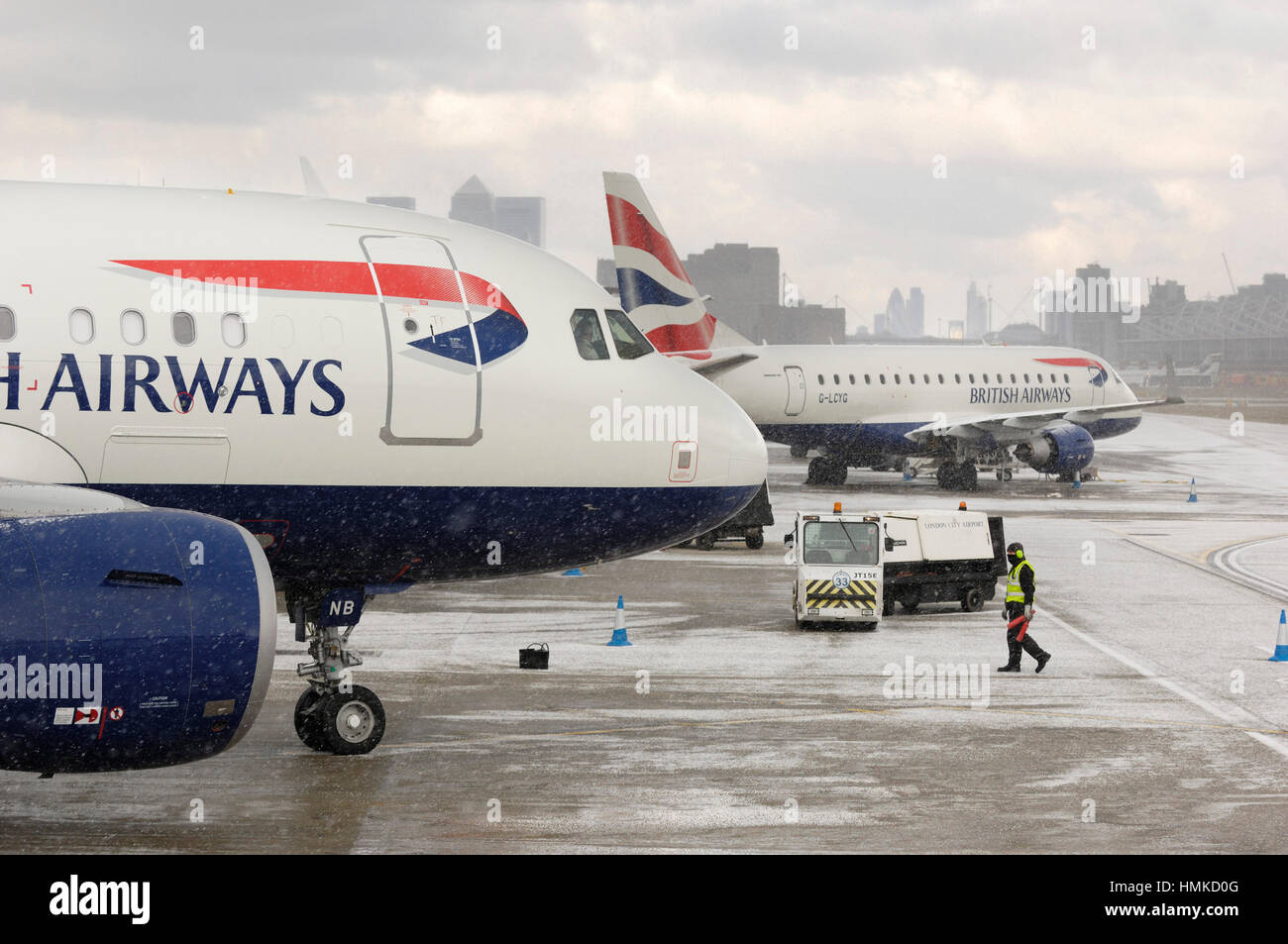 marshaller wearing yellow high viz tabard ear-defenders standing British Airways BAW Airbus A318-100s parked in snow Stock Photo