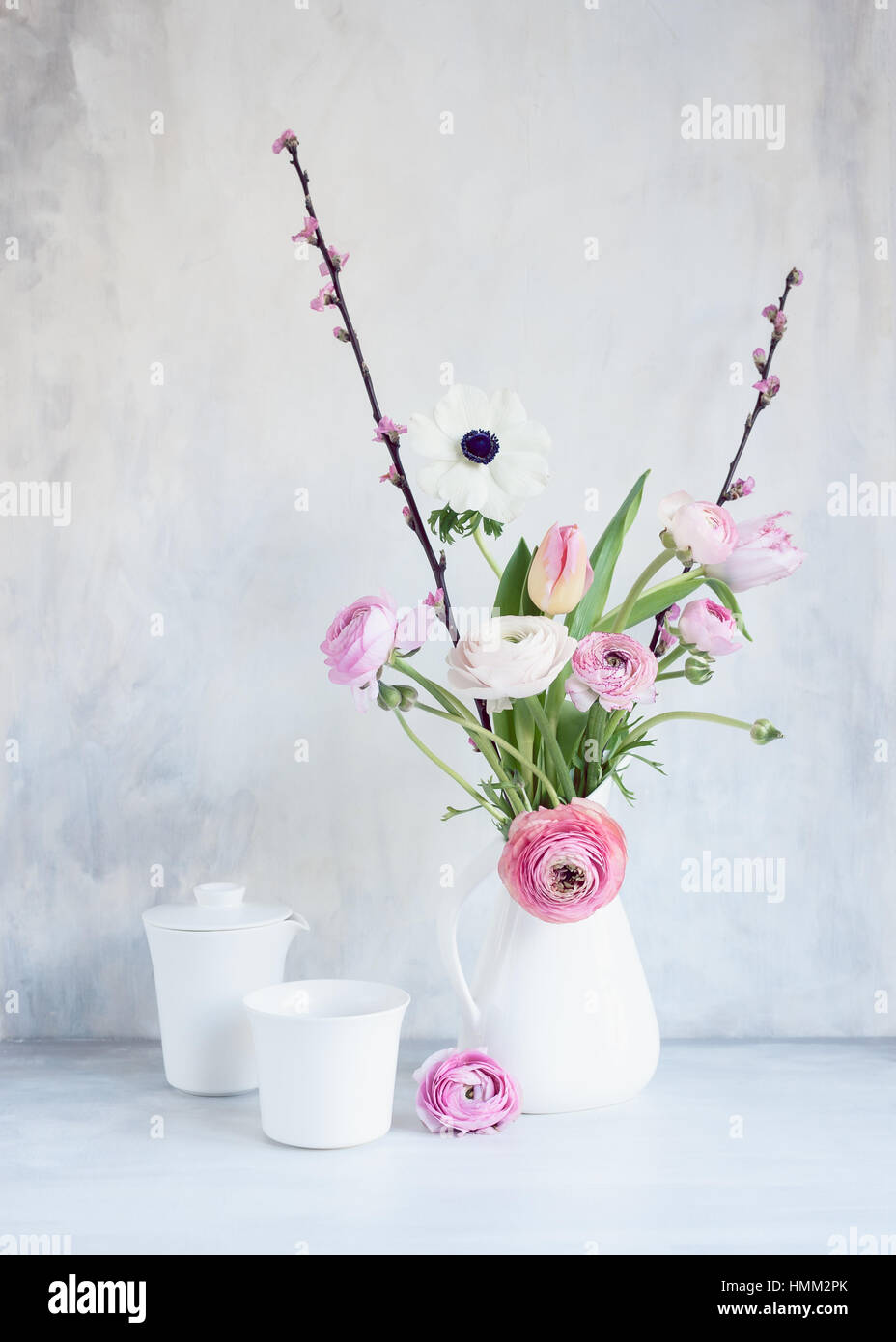 Still life of pink white spring flowers in white jug with cup and teapot, on a table, against painted backdrop Stock Photo