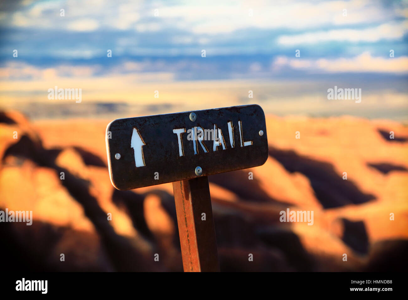 A rustic trail sign looms over a scenic view in Arches National Park in Utah. A white arrow points in the direction the traveler should go. Stock Photo