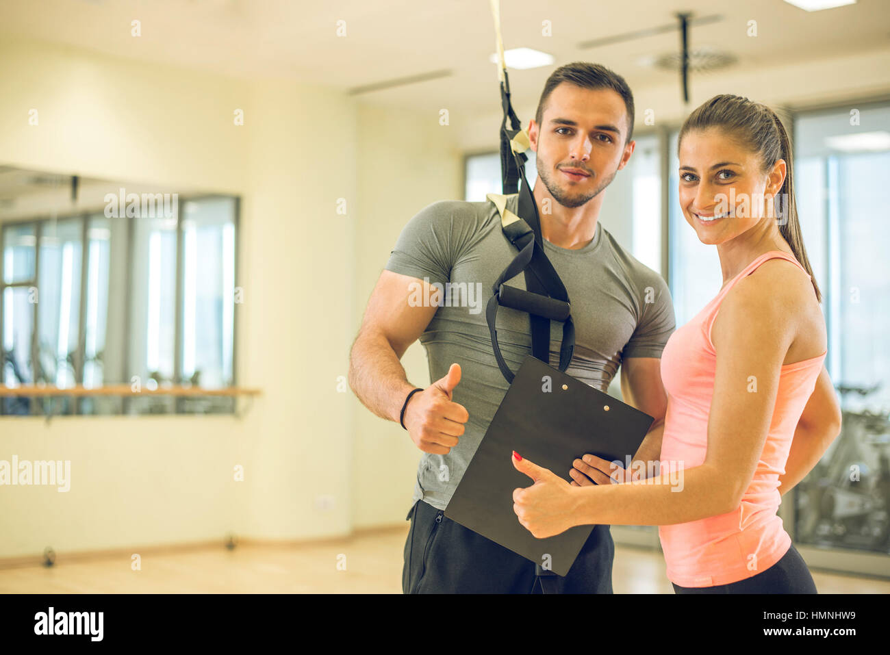 Personal trainer showing result of training plan to his female young client with suspension rope over shoulder. Stock Photo