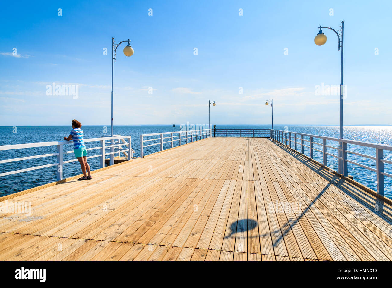 Wooden pier and young woman standing against railing in distance in Jurata town on coast of Baltic Sea, Poland Stock Photo