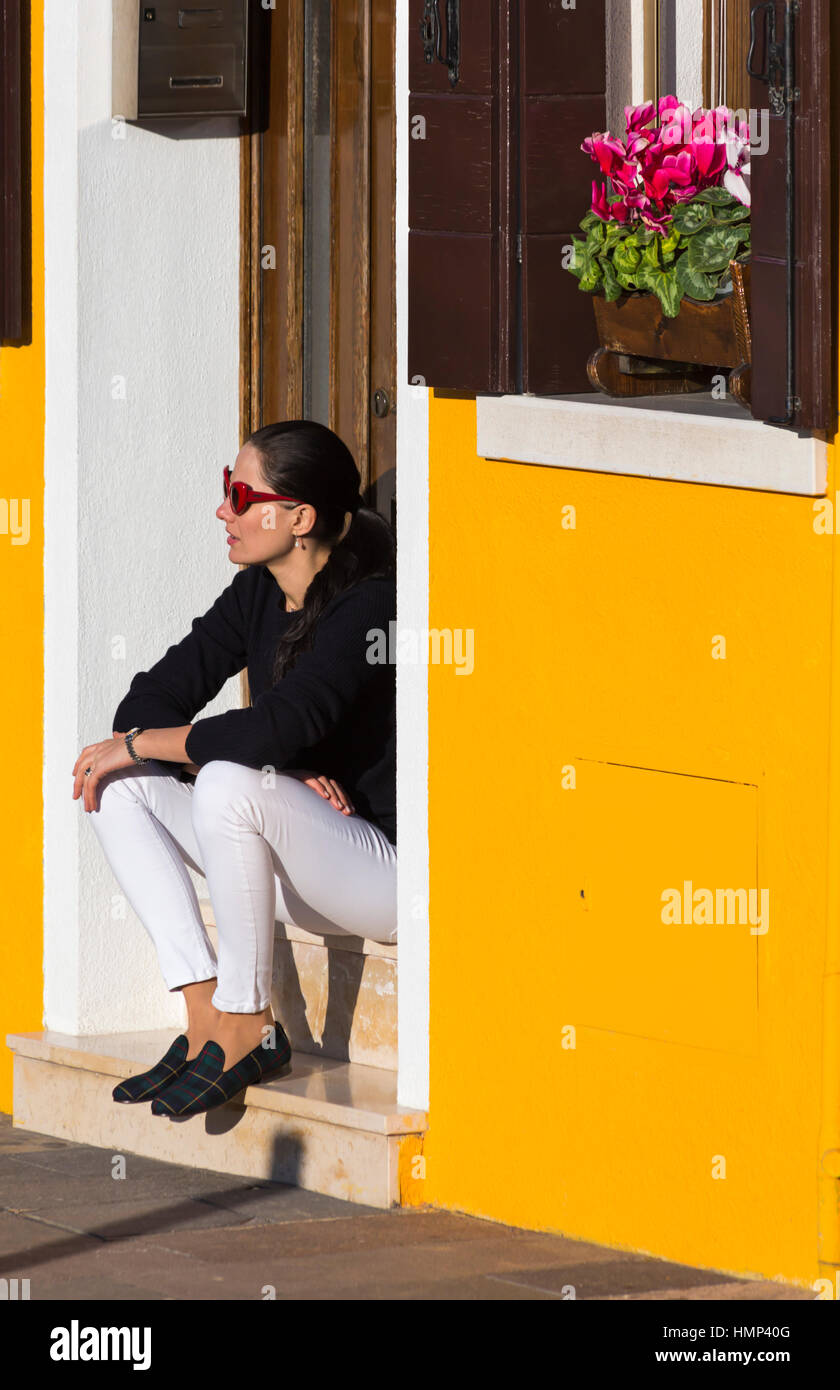 Woman sitting on steps in doorway of brightly coloured yellow house at Burano, Venice, Italy in January Stock Photo