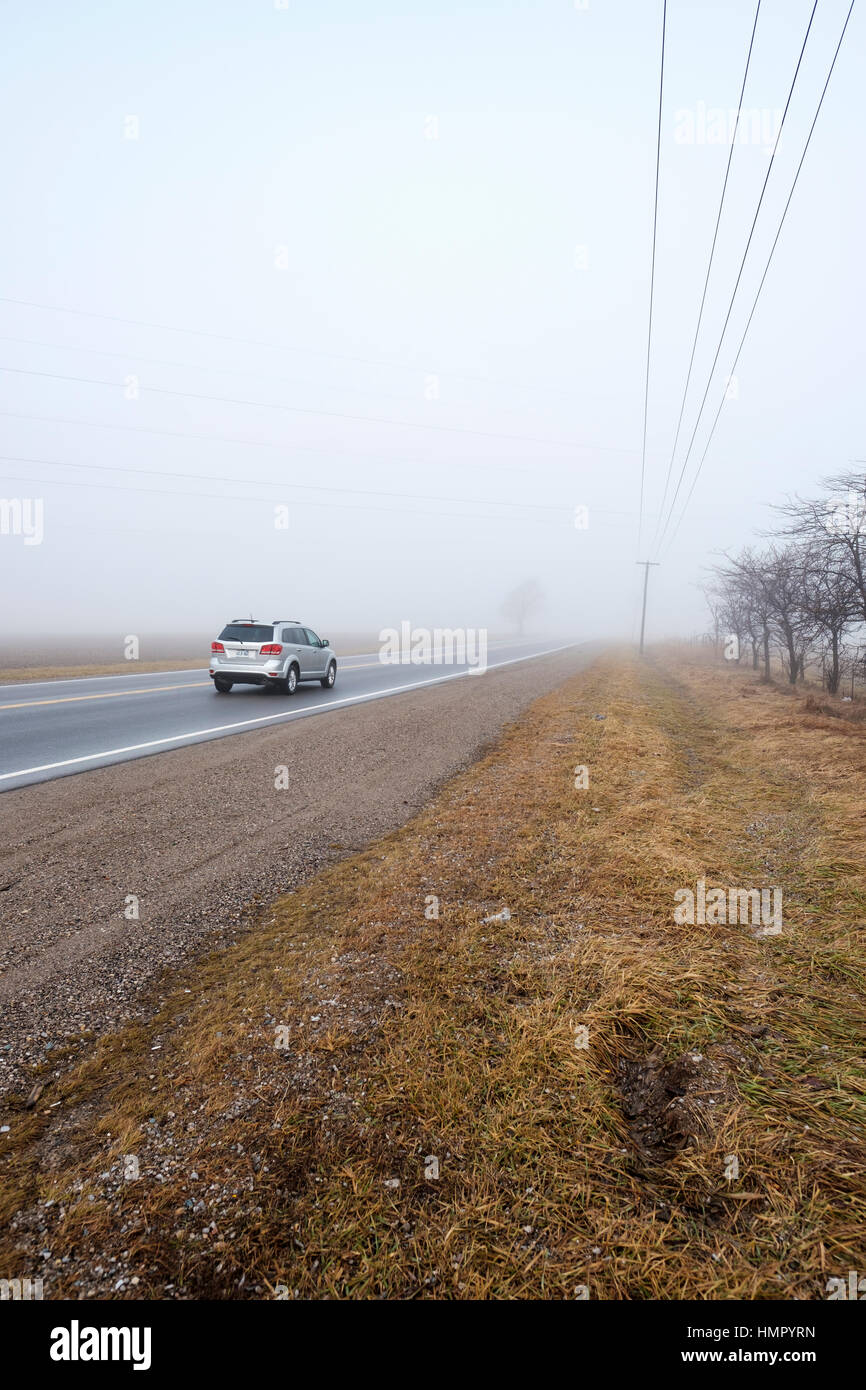 A silver vehicle drives on a paved rural road in a foggy morning in Southwest Ontario, Canada. Stock Photo