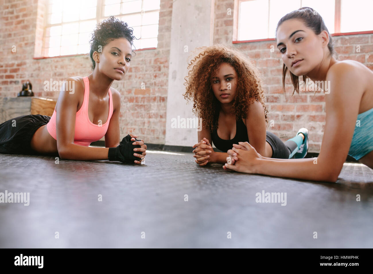 Portrait of three young women lying on gym floor and looking at camera. Females in fitness class. Stock Photo