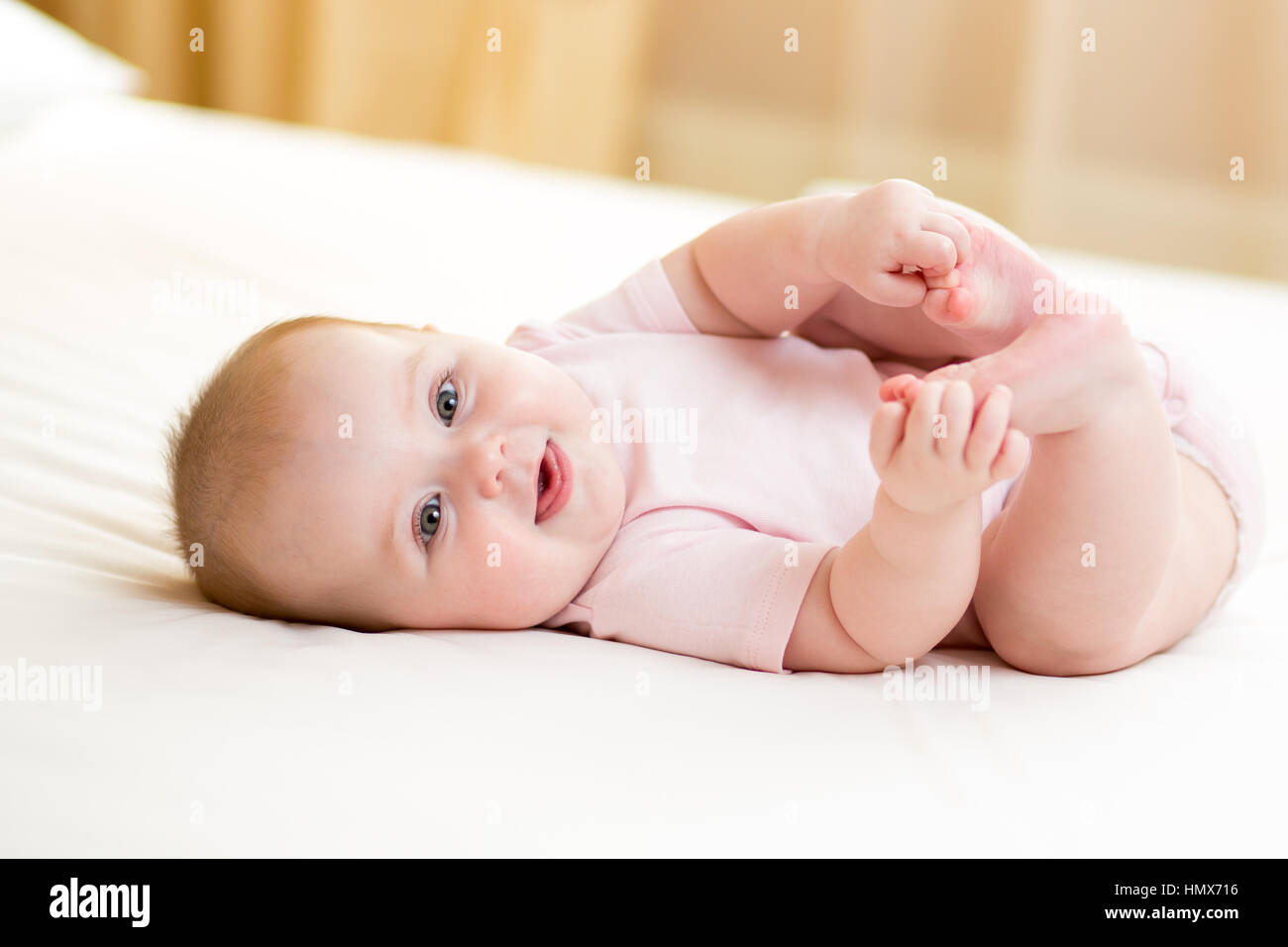 happy baby girl lying on white sheet and holding her legs Stock Photo