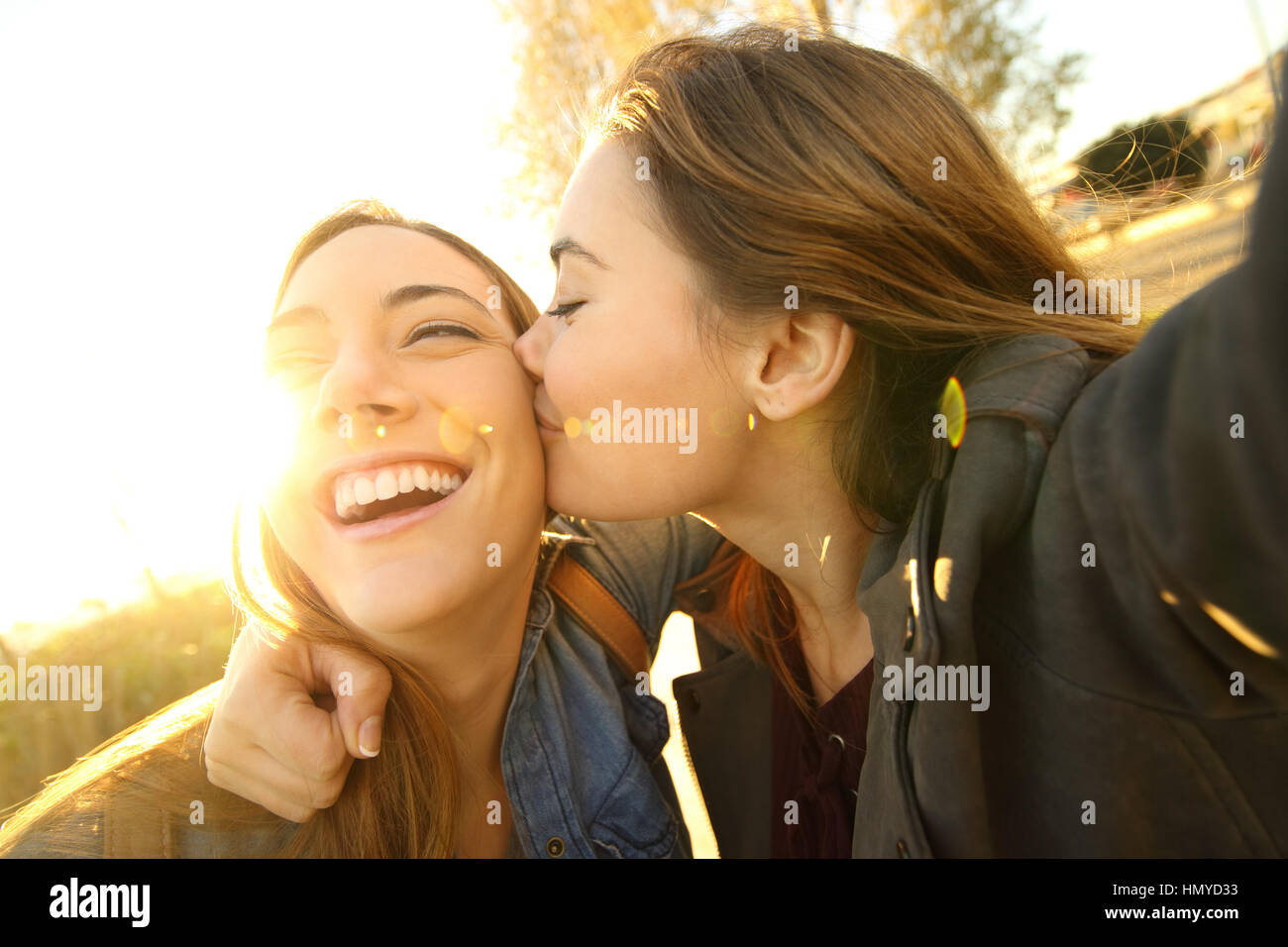 Two affectionate friends kissing and taking a selfie outdoors in the street at sunset with a warm light in the background Stock Photo