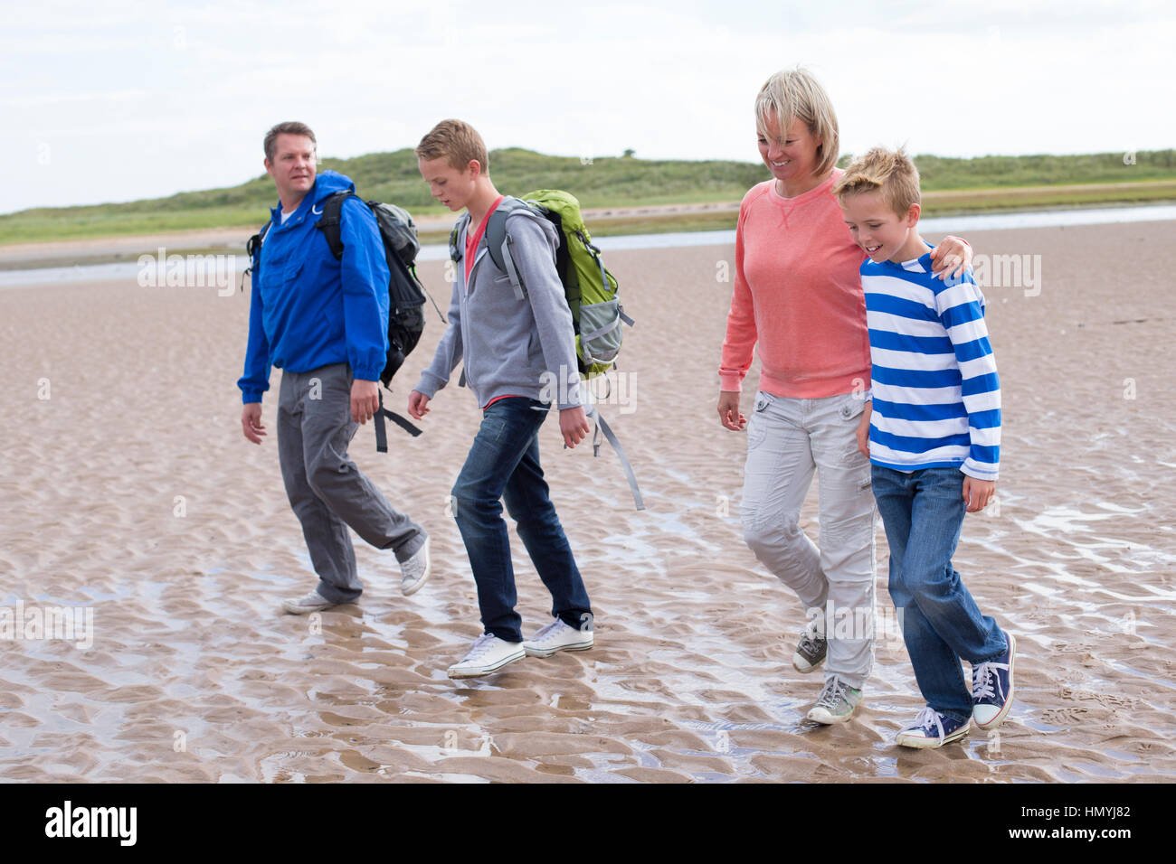 Family of four walking across the beach. They are wearing warm casual clothing with backpacks and look very happy. Stock Photo