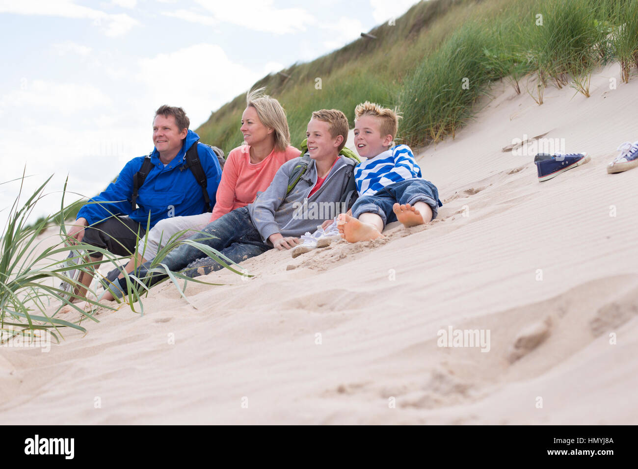 Family of four sitting on the sand dunes. They are wearing warm, casual clothing and looking out to sea. Stock Photo