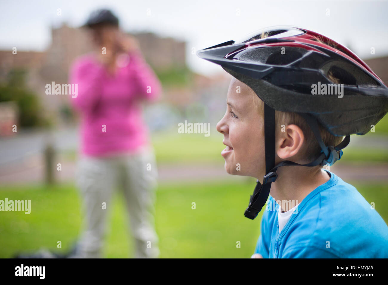 Child in the foreground with a bicycle helmet on while his mother is in the background taking a picture of him. Stock Photo