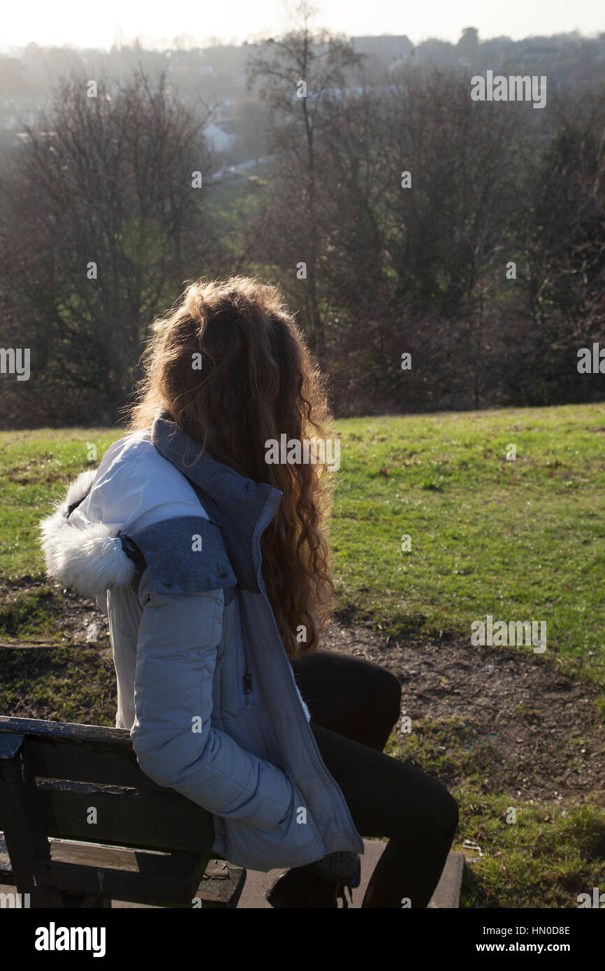 Young female sitting  on a bench, back to camera. In a park open space. Stock Photo