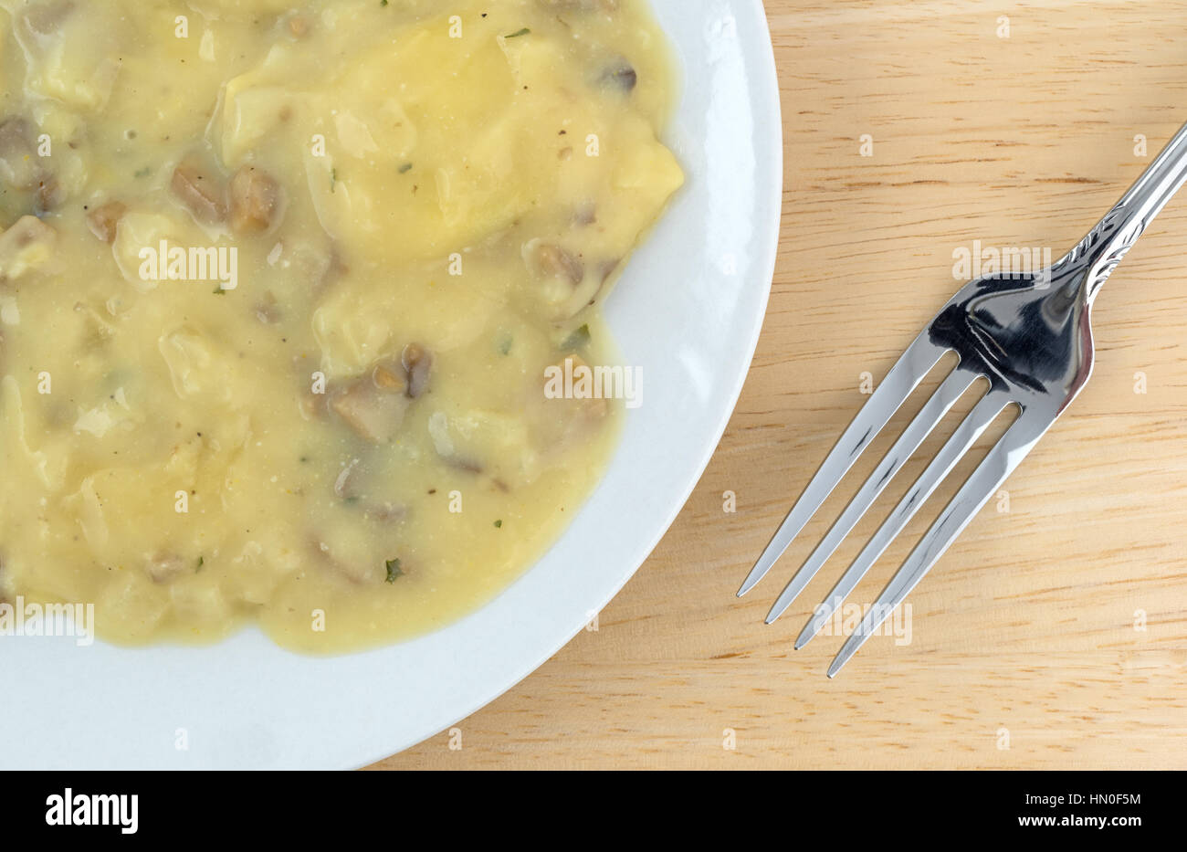 Top close view of a ravioli covered with a cheese and mushroom sauce meal on a plate with a fork to the side on a wood table top. Stock Photo