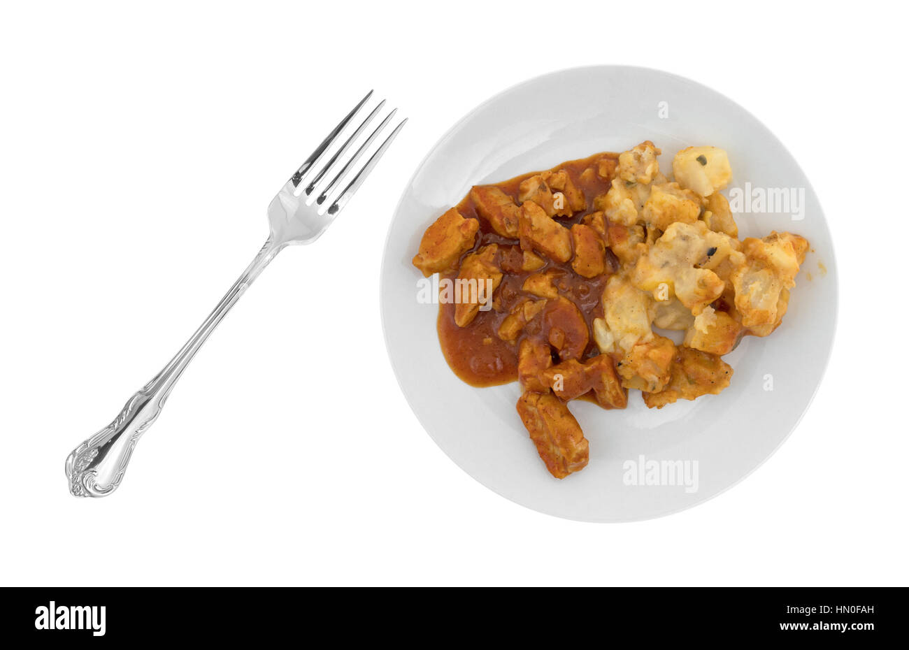 A plate with chunks of chicken in a thick barbecue sauce plus cheese covered potatoes plus a fork to the side isolated on a white background. Stock Photo