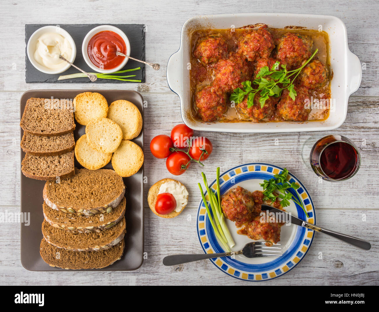 Delicious meal rich in protein and vitamins. Meatballs with tomato sauce, tomatoes, bread, mayonnaise, ketchup and a glass of wine on a white wooden b Stock Photo