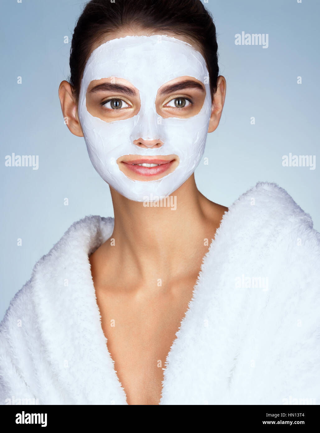 Smiling woman with moisturizing facial mask. Photo of brunette woman wearing white bathrobe. Beauty & Skin care concept Stock Photo