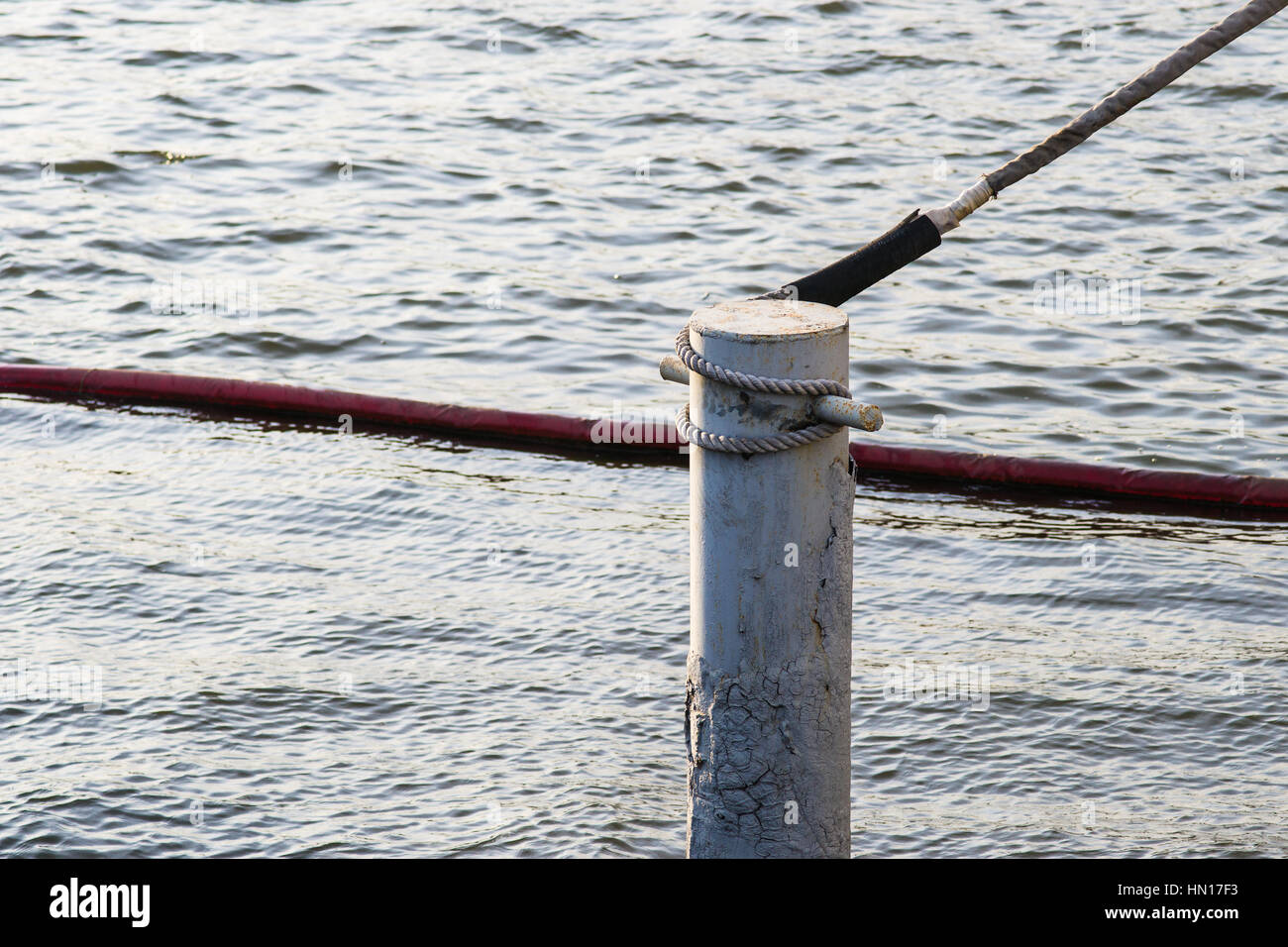Water of the river, metal pole or riding bitts, anchor tow tied to the pole. Red cable lies in the water. Minimalistic industrial composition. Stock Photo