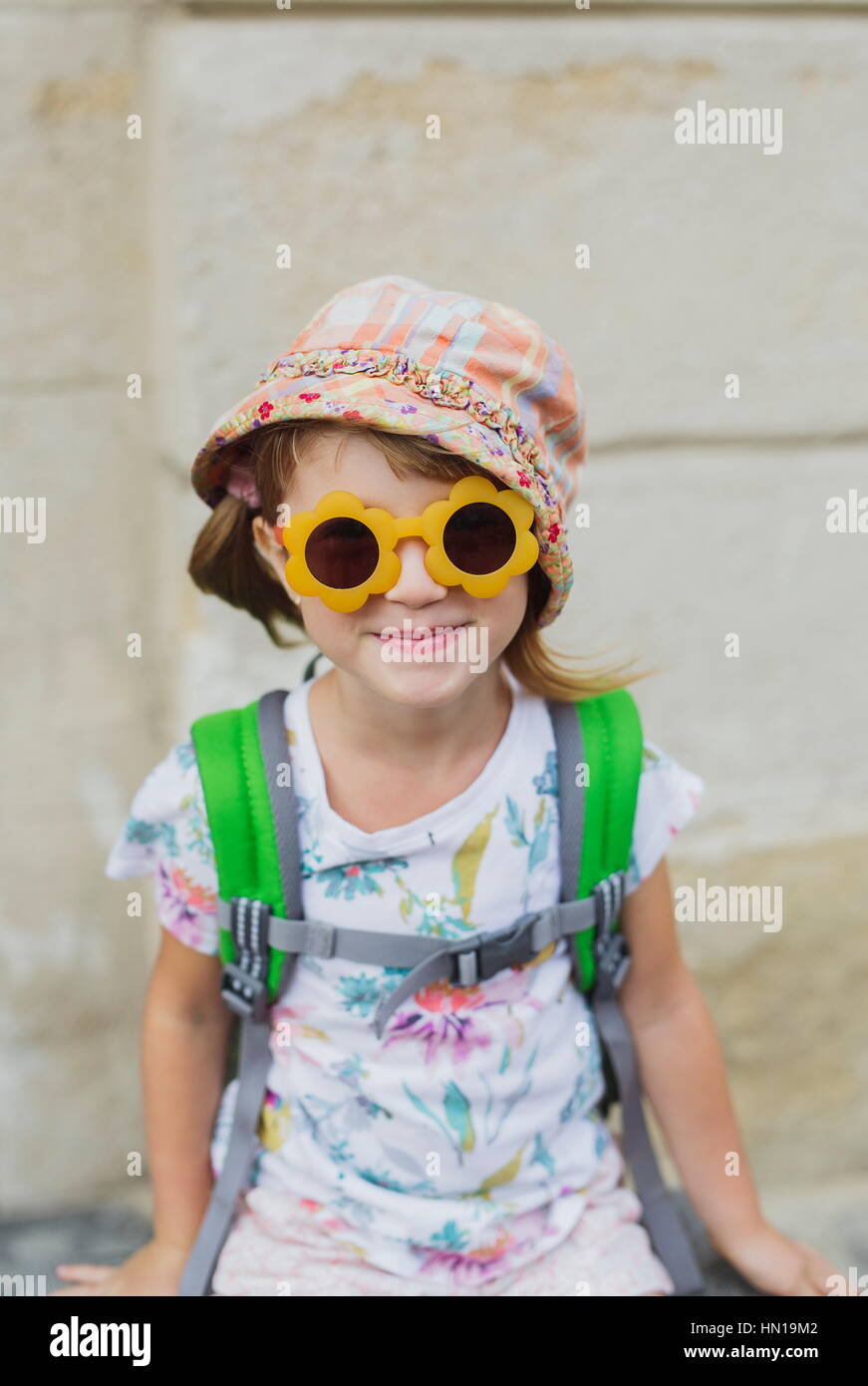 Cute smiling happy little girl with a backpack sitting on a bench. Child tourist in big old city. Kid in sunglasses during summer vacation. Happy litt Stock Photo