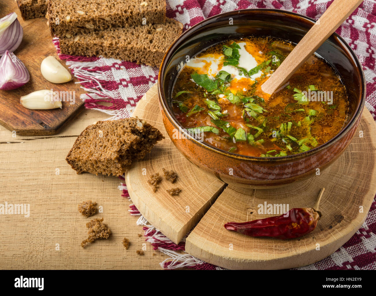 Tasty and nutritious soup with meat, potatoes, cabbage, tomatoes, beans, sour cream, parsley, onion, garlic, dark bread in a clay plate on a wooden tr Stock Photo