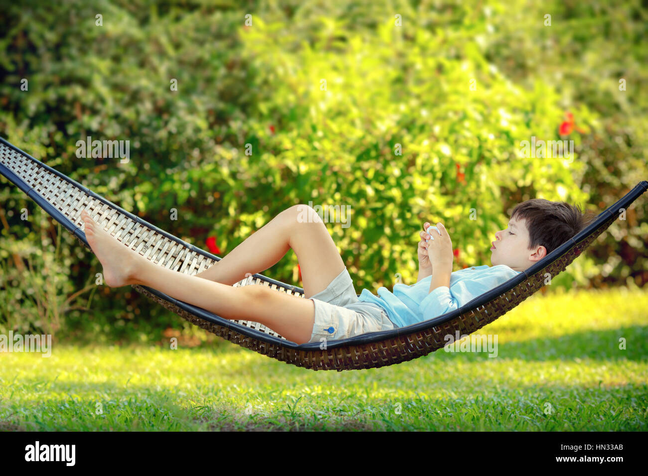 Side view of cute little boy lying on a hammock looking at his cell phone and relaxing Stock Photo