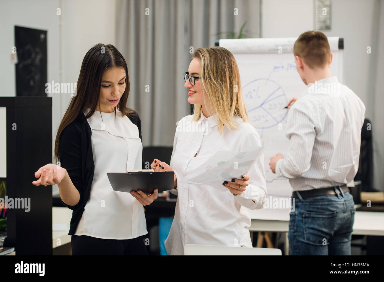 Two beautiful office workers having conversation while man colleague drawing business strategy on flip chart Stock Photo