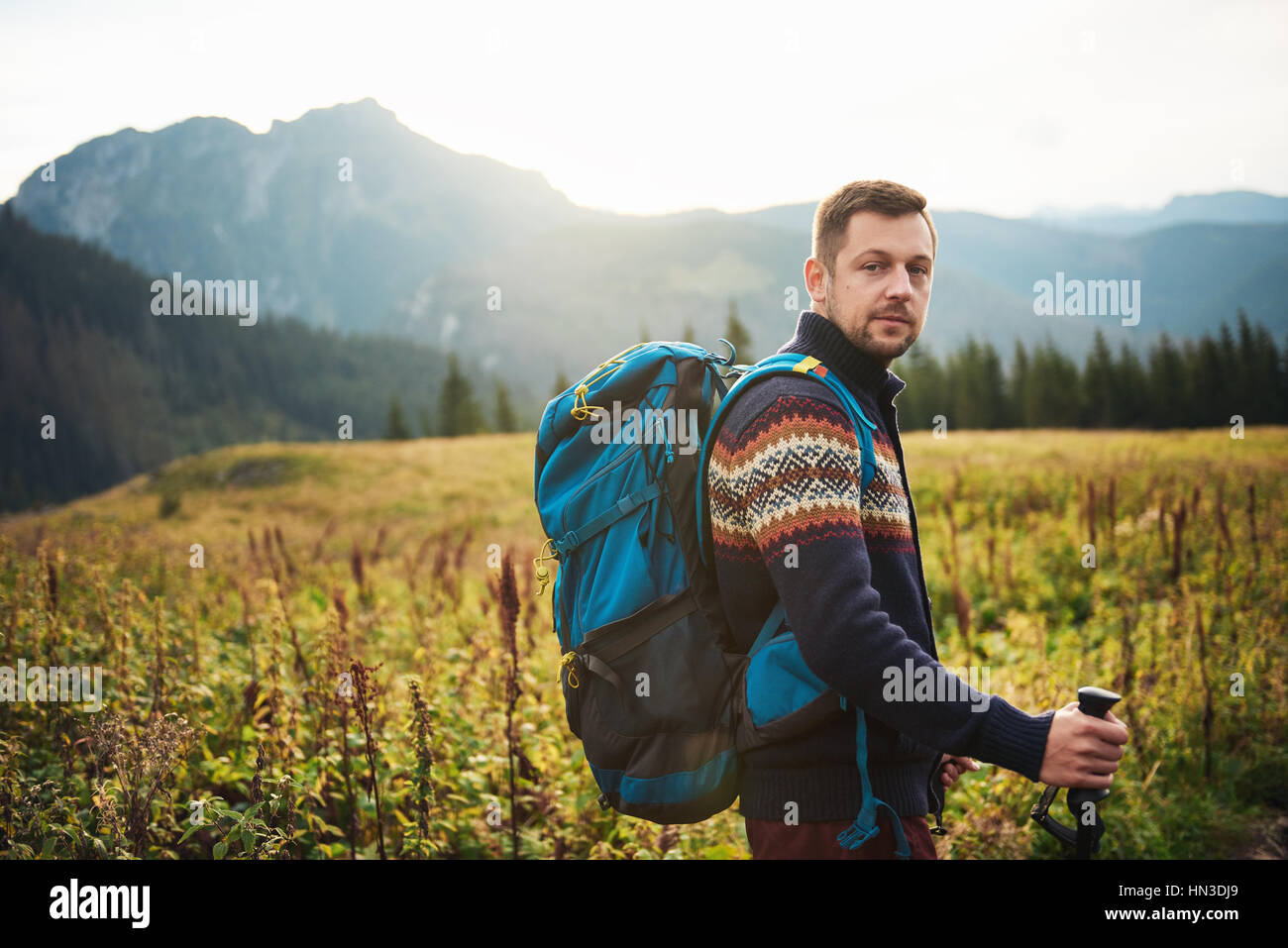 Portrait of a young man wearing a backpack and carrying trekking poles standing in a field while hiking in the wilderness Stock Photo