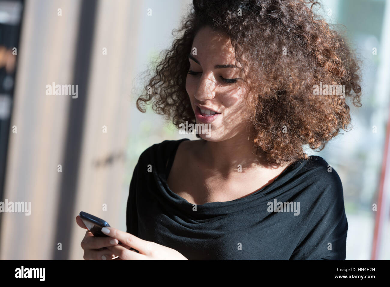 Attractive woman with lovely curly brown hair lit by the sunlight standing reading a text message on her mobile with a wry smile Stock Photo