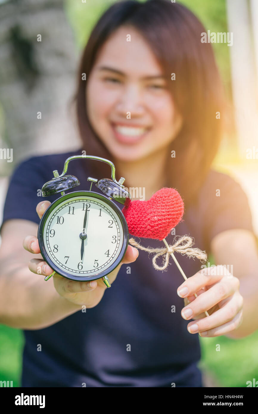 love and time or time for love concept, happy smile Thai Asian cute teen girl woman hold clock and red heart in green park background. Stock Photo