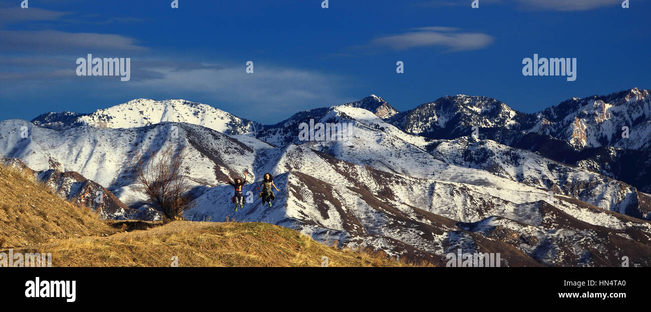 Two young girls jump on a hiking trail overlooking the Wasatch mountains of Salt Lake City, Utah. The mountain peaks are snow-covered and dramatic. Stock Photo