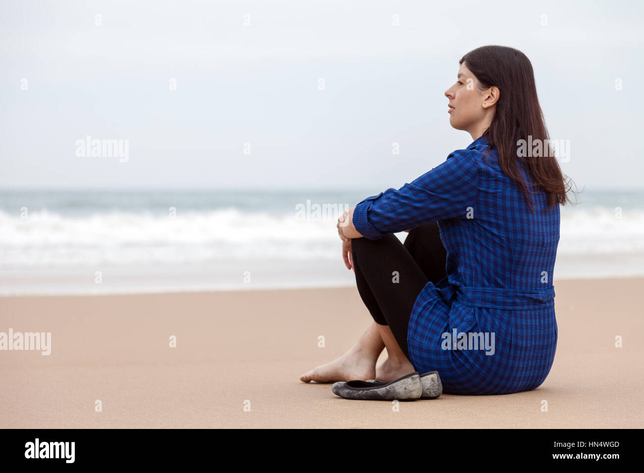 Lonely and depressed woman sitting on the sand of a deserted beach and watching the sea on an Autumn day/ woman beach alone lonely sitting sad sadness Stock Photo