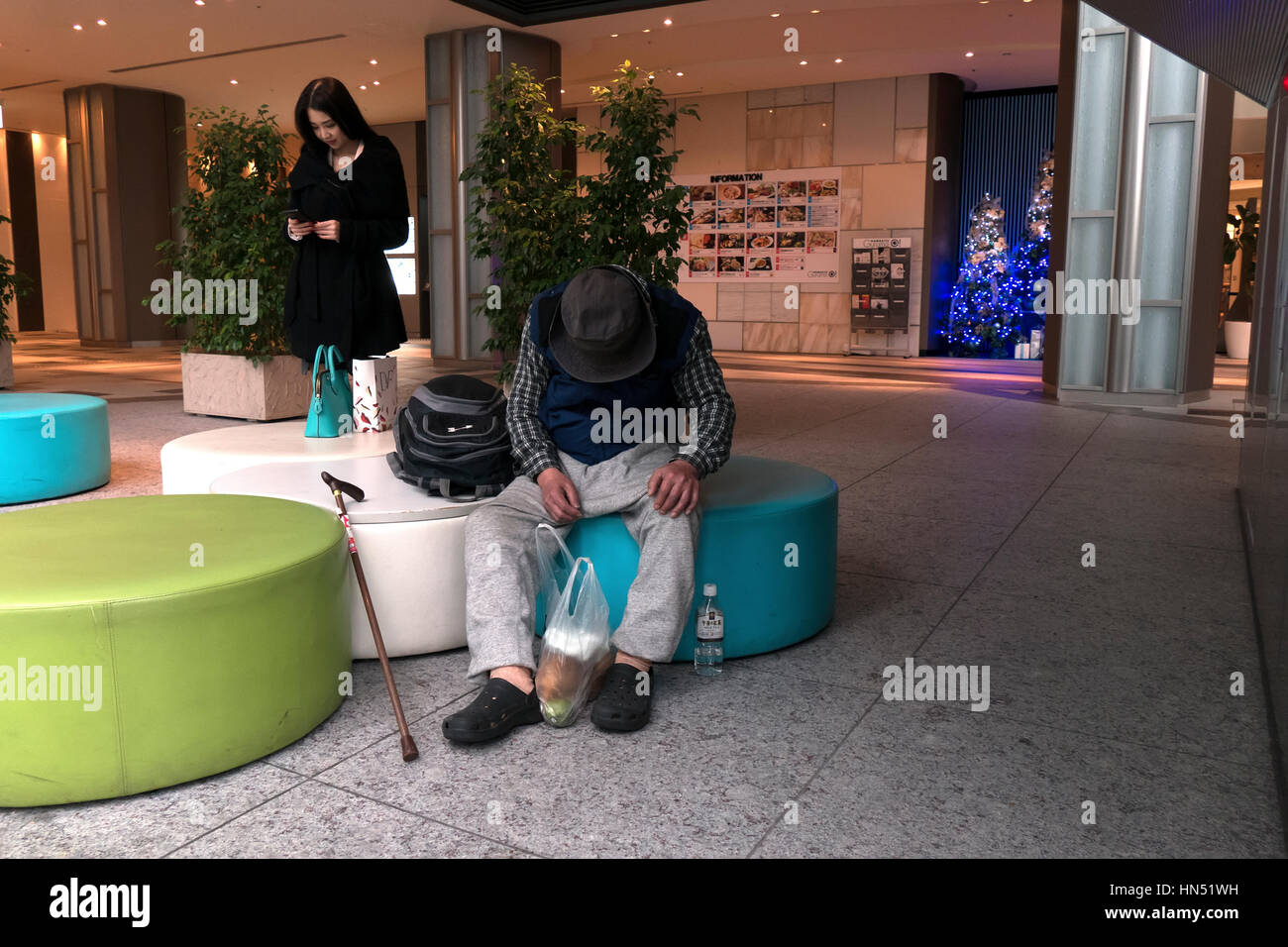 Beautiful Japanese woman texting on smartphone and homeless man sleeping in the lobby of a modern building. Tokyo, Japan, Asia Stock Photo