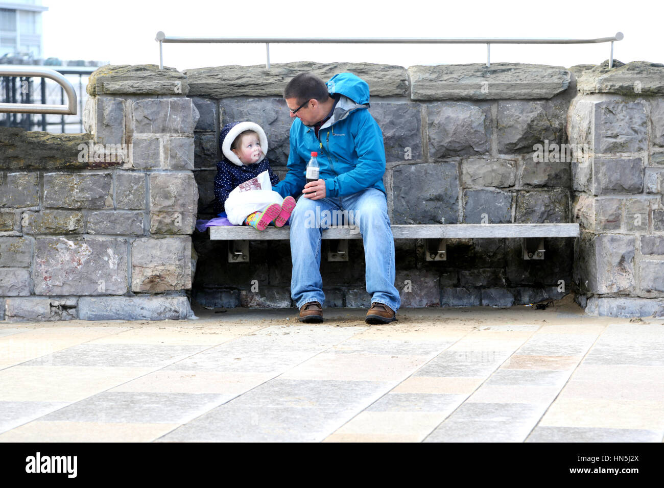 A grandfather sitting outdoors on a wooden bench sharing a bag of traditional fish and chips with his young granddaughter Stock Photo