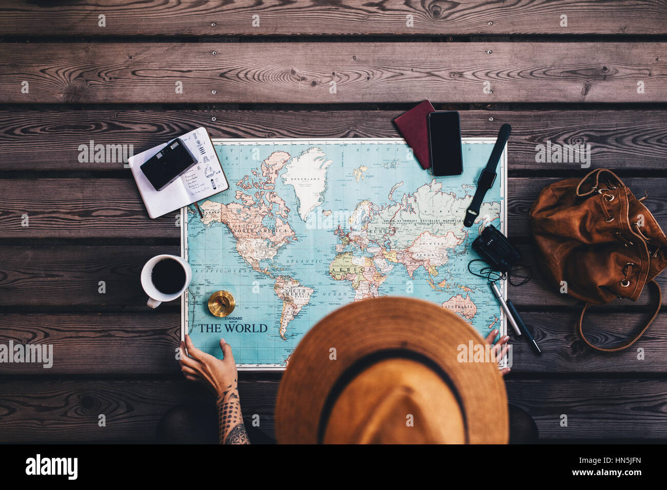Young woman planning vacation using world map and compass along with other travel accessories. Tourist wearing brown hat looking at the world map. Stock Photo