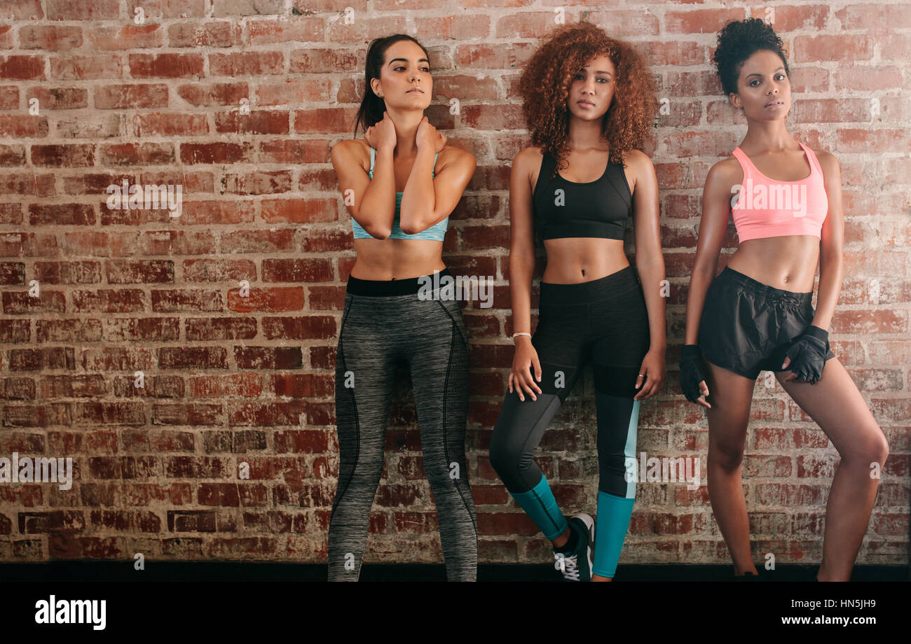 Portrait of three fitness women standing against brick wall in gym. Multiracial group of females in sportswear standing together. Stock Photo