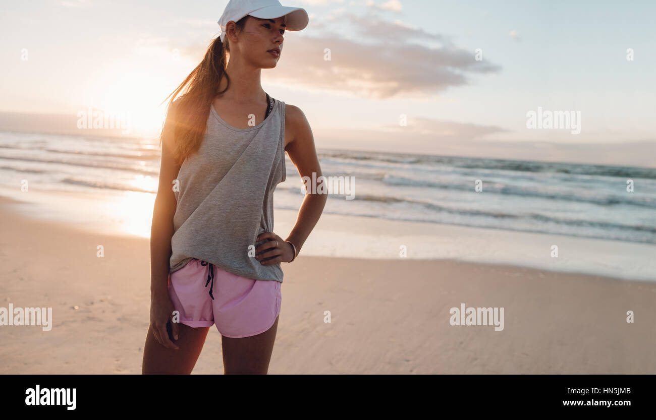 Shot of healthy young woman standing on the beach with her hand on hip and looking away. Female model in sportswear on the sea shore in morning. Stock Photo