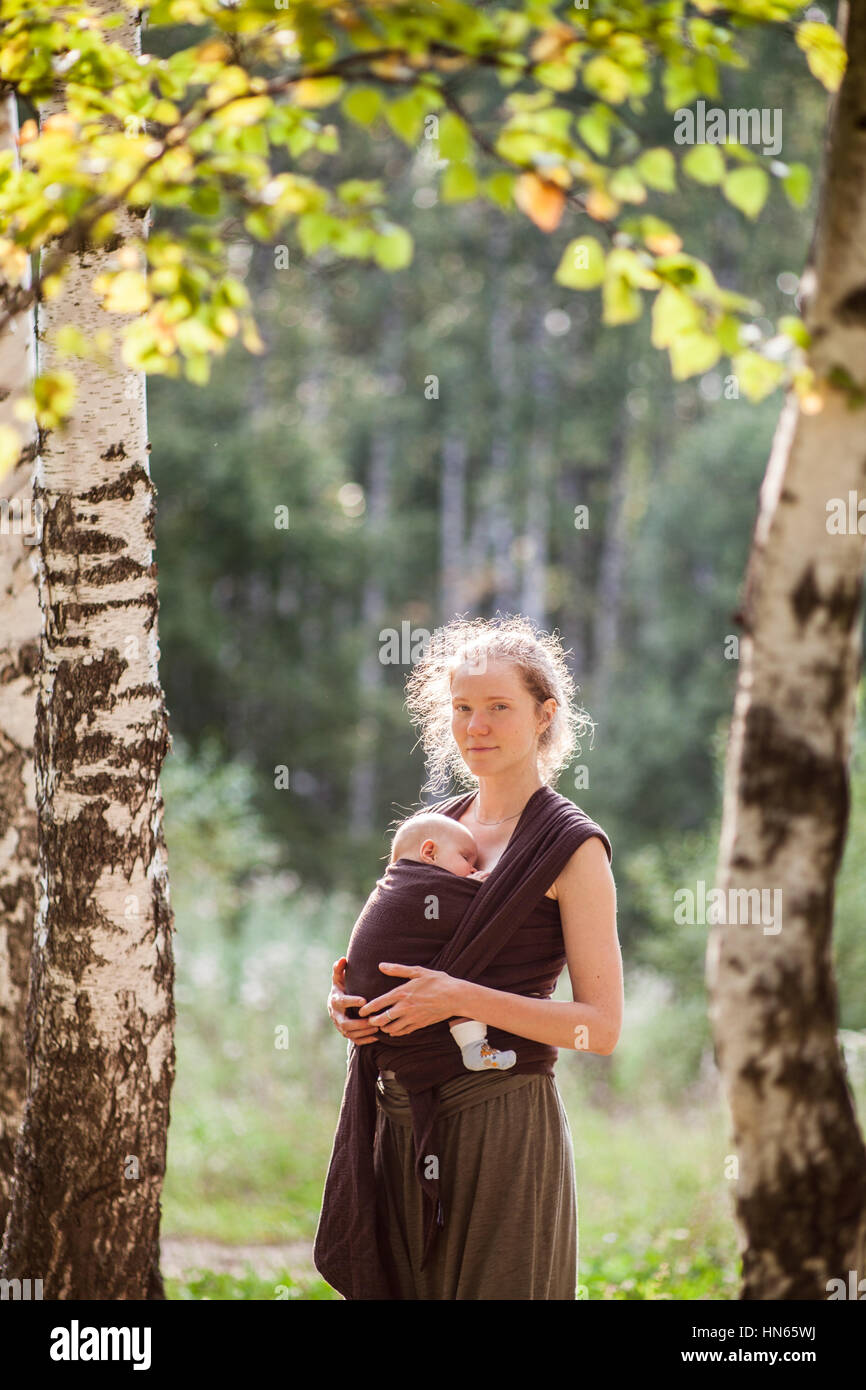 Mother carrying her child in a baby sling scarf in the sunny forest Stock Photo