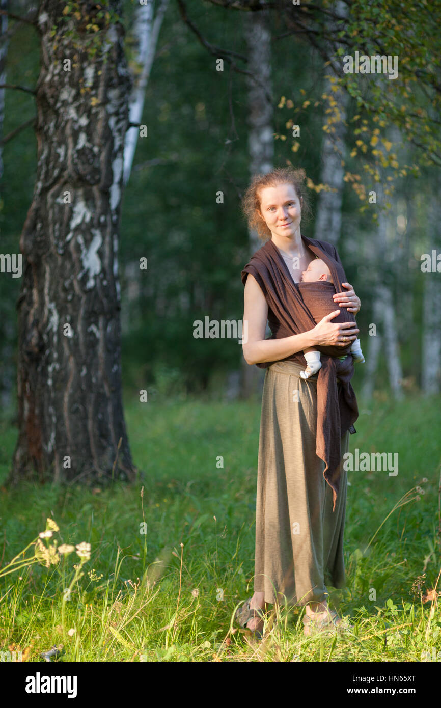 Mother carrying her child in a baby sling scarf in the sunny forest Stock Photo