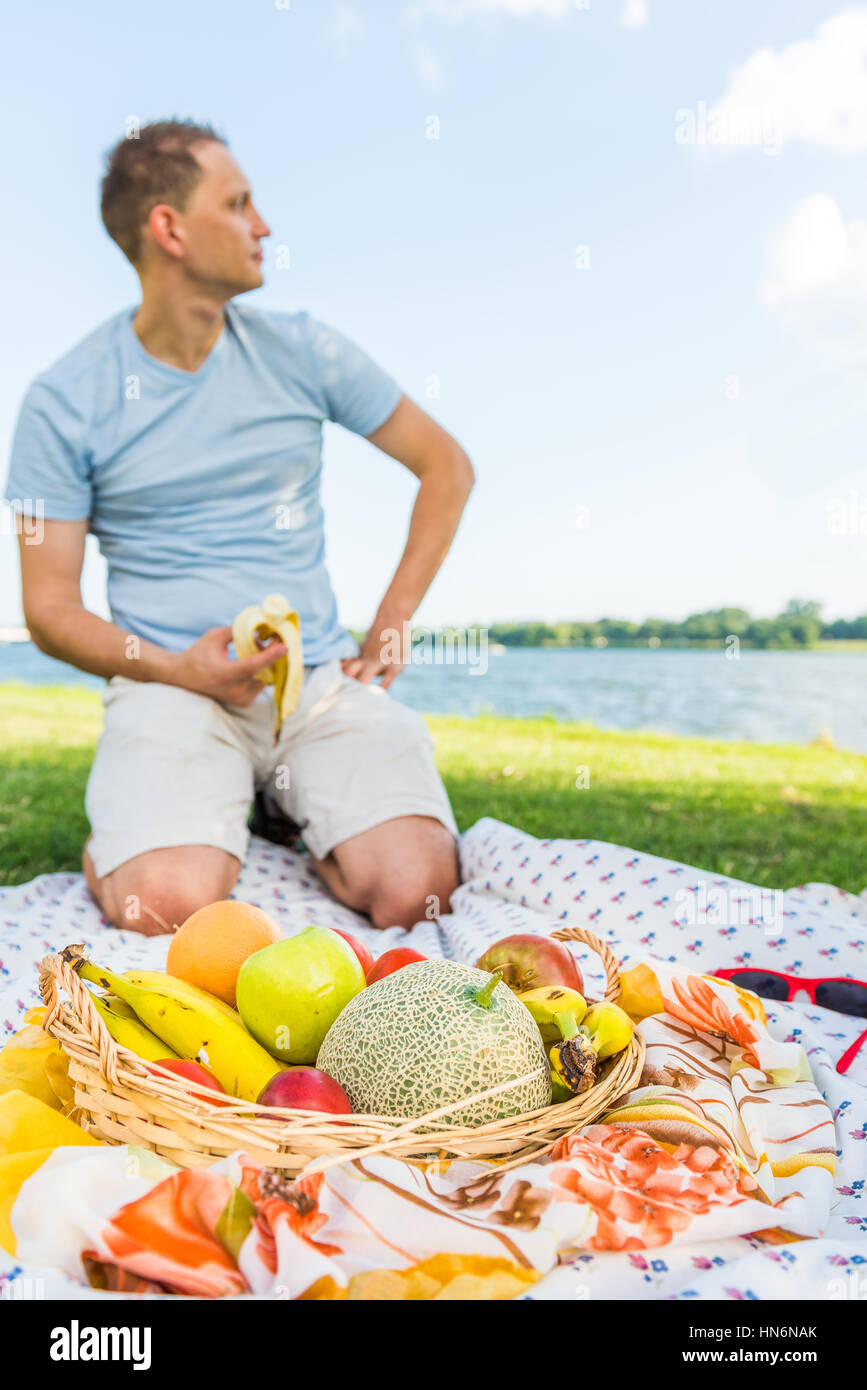 Young man sitting on picnic blanket eating banana by fruit basket looking at river Stock Photo