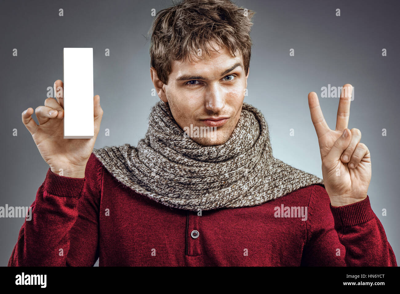 Man holding white box of medicine and shows two fingers. Photo of unhealthy man wrapped scarf. Health care concept Stock Photo