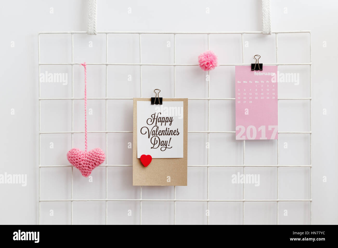 Happy Valentine's Day card on a metal grid display with knitted pink heart and pom pom. Valentine's Day decoration. Wall grid organizer. Stock Photo