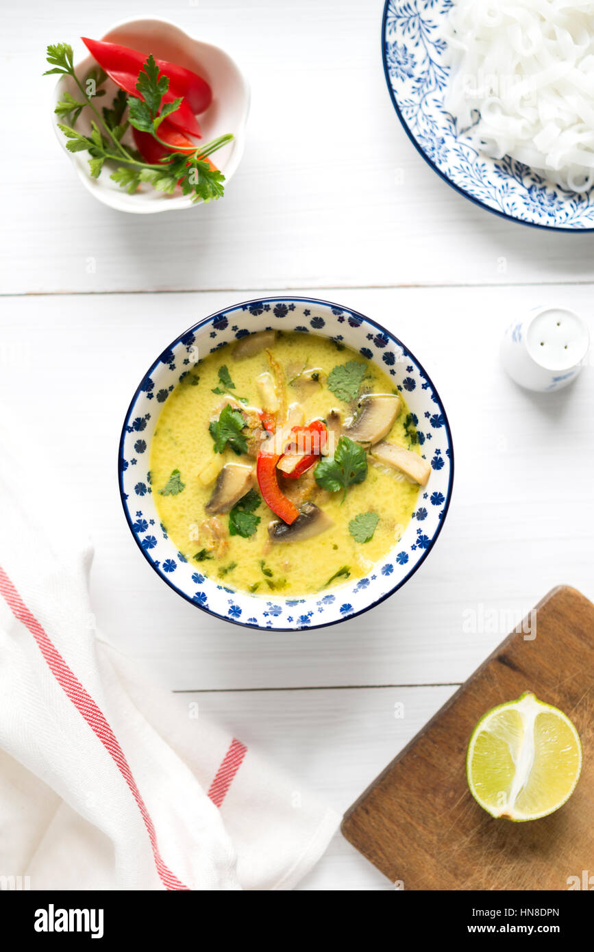 Thai coconut milk soup with chicken meat, mushroom, coriander, red pepper and rice noodles in colorful bowl on white table. Top view. Stock Photo