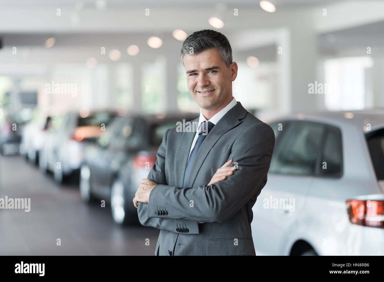 Confident smiling car salesman at the showroom, he is standing with arms crossed, luxury cars on the background Stock Photo