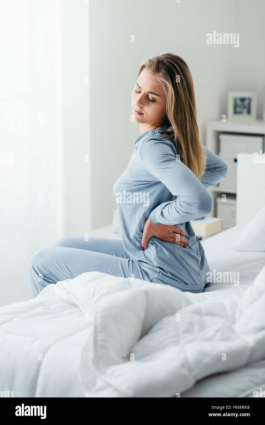 Young attractive woman with back pain waking up and sitting on her bed